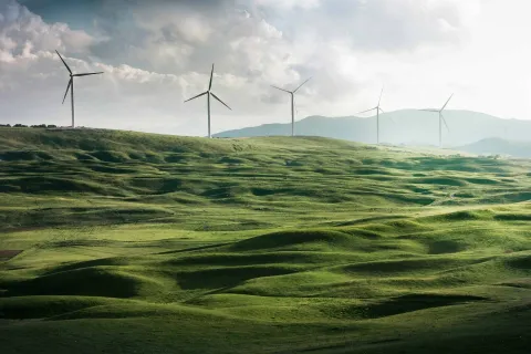 Five wind turbines in the distance on green hills, with mountains and clouds in the background.