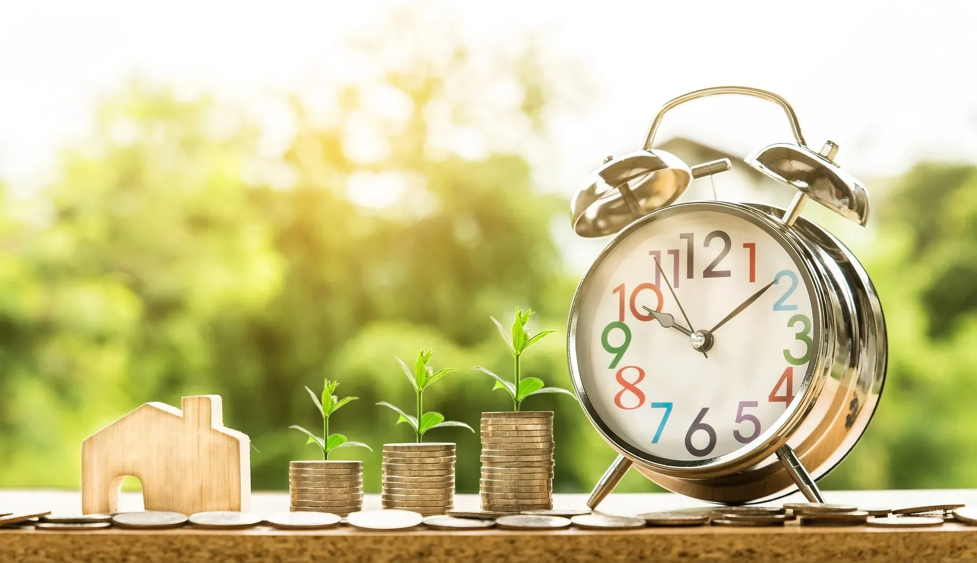 An alarm clock sits next to three neatly stacked piles of coins and a wooden block carved into the shape of a house.